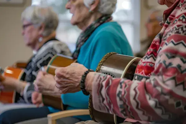 Three older adults sit side by side, playing hand drums, as part of a small group music session or recreational activity in a community or senior center.