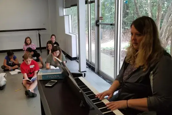 An individual plays an electric keyboard at the front of a bright room while several children sit on the floor in the background near large windows.