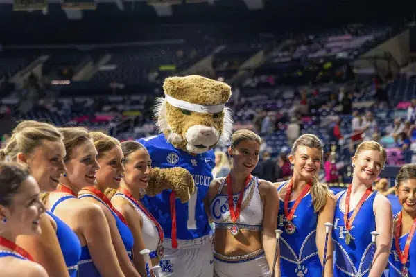 The nine-member team wearing their UK blue uniform with medals around their neck poses for a photo with the Wildcat mascot in front of a crowd.
