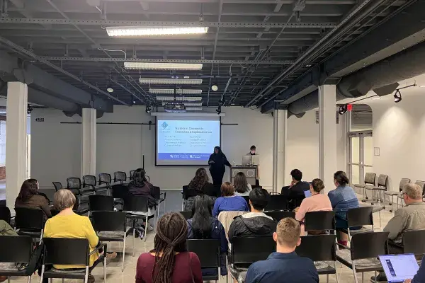 A speaker presents to a seated audience in a loft-style event space with exposed ceiling ductwork. A projection screen displays a slide titled 'Hardware, Community Connection & Implementation' with presenter names Paula Farrell and Rupin Sharma, both from the Department of Pediatrics, College of Medicine, alongside logos for UK College of Fine Arts, SA/VS, and the Office of the Vice President for Research. A presenter stands near a podium with a laptop, addressing about 20 attendees seated in rows of black 