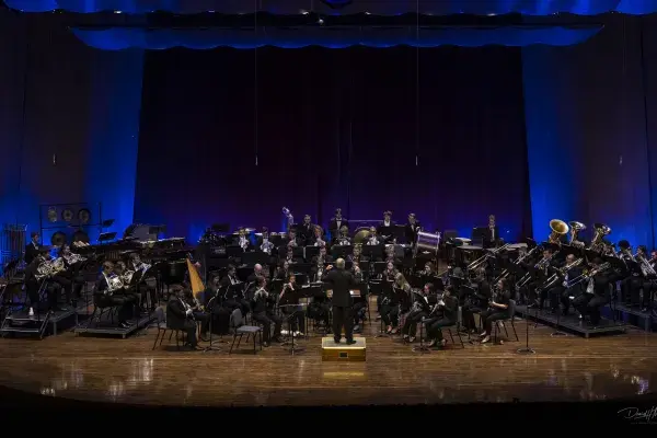 Photo of UK Bands performing onstage at the Singletary Center with instruments, in concert blacks, conductor facing away from camera leads the musicians, with blue uplighting behind them