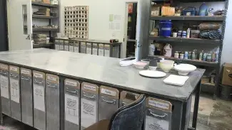 A large high metal table takes up the center of the room with vertical drawers below labled for various glaze and pigment materials. A tile color swatch board is on the far back wall and several shelves with more supplies.