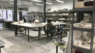 A brightly lit room lined with shelves for projects and supplies. Several large tables are arranged in the center of the room as communal workspace with many stools available.