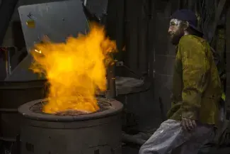 A student stands back from a burning furnace. A pillar of red and gold flame blooms out of the top of the furnace.
