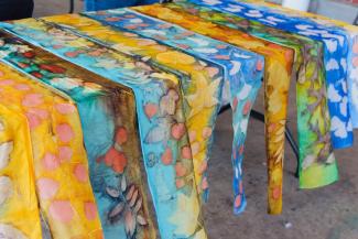 A row of bright multicolor silk scarves with arious leaf and twig patterns laid out across a table to dry.