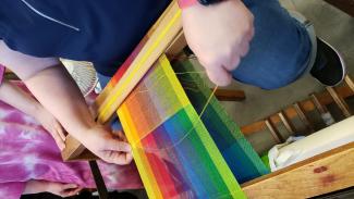 A rainbow of thread colors is warped onto a floor loom while a student works at weaving in a bright yellow weft.