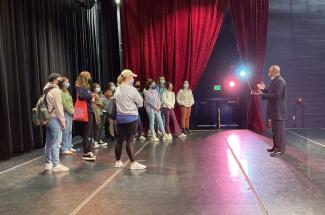 Students onstage at the Lyric listen to Whit Whitaker, wearing a black suit
