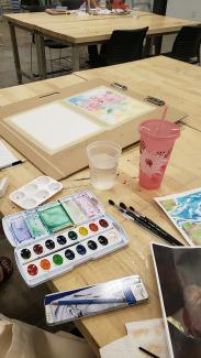 A table with a watercolor palatte open, several brushes, some references images to the side, and a painting in progress.