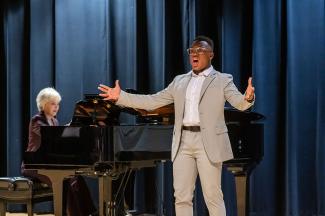 CoryOn Brooks (2024) performing in front of a dark blue velvet curtain, with Professor Nan McSwain accompanying on a grand piano in the background. Photo by Sally Horowitz Photography.