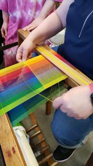A rainbow of thread colors is warped onto a floor loom while a student works at weaving in a bright yellow weft.