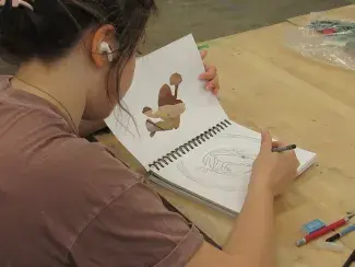 Viewed from over a student's shoulder as they listen to music and sketch a frog illustration in a notebook. Other pencils and block erasers sit nearby on the table.