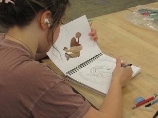 Viewed from over a student's shoulder as they listen to music and sketch a frog illustration in a notebook. Other pencils and block erasers sit nearby on the table.