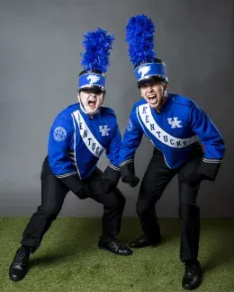 two students in blue university of kentucky band uniforms with mouths open shouting in excitement