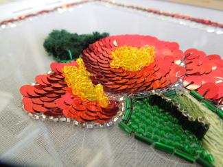 A thin white netting fabric is stretched over a frame and adorned with bright red sequins, yellow glass beads, and metallic green beads to form the shape of poppies with leaves.