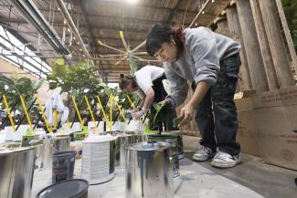 student bends down to dip a paintbrush in one of many paint cans on the ground
