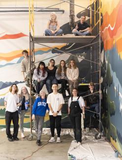 group of students pose in front of large mural in progress on building wall