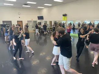 View from a high vantage point of a brightly lit dance studio full of people sectioned off in group of two to three moving to music. The mirrors along the back wall reflect the dancing students as well as the scenery outside large windows on the opposite side.