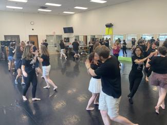 View from a high vantage point of a brightly lit dance studio full of people sectioned off in group of two to three moving to music. The mirrors along the back wall reflect the dancing students as well as the scenery outside large windows on the opposite side.