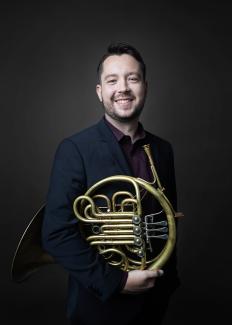 Portrait of Ryan Little in a dark studio with a navy blue suit holding his bright horn.