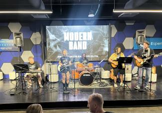 Five performers on a stage (left to right: man on keyboards, woman on electric guitar, woman on drums, person on electric mandolin, man on traditional guitar with pickups) play in front of a blue, white, and black hexagon pattern with "Modern Band" projected behind them.
