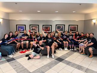 A class of 17 smiling students wearing matching t-shirts sit around a large couch area in the Guignol Theatre lobby. An assortment of performance photos hang on the wall behind them. Each of the students is holding one end of a colored ribbon in each hand while the ribbons criss-cross and twist around the group where other students to hold the opposite end.