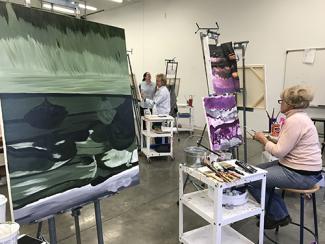 A studio with multiple paintings in progress on easels, some being worked on by students. The abstract painting in the foreground is done entirely in broad strokes of shades of green. Another student works in the background on a pink and purple landscape.