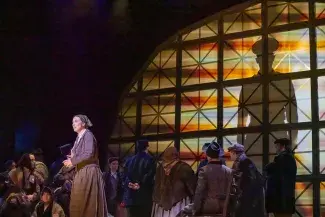 2026 production of A Nation of Others. A spotlight shines on a worried immigrant woman in drab early 1900s clothing, a crowd mills in the background. Behind all of them on a massive window is cast the silhouette of a watchful police officer.