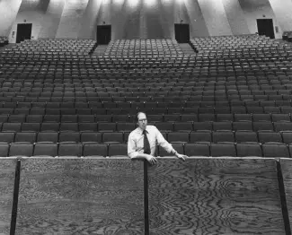 Black and white photo. Dean Wills stands in the front row of the brand new Center for the Arts at UK with rows of seats stretching up toward the back wall far behind him.