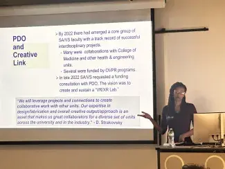 A presenter stands at a lectern and references a powerpoint presentation projected on a screen behind. 