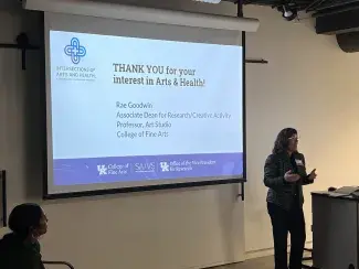 College of Fine Arts Associate Dean for Research/Creative Activity Rae Goodwin stands in front of a projector screen that welcomes attendees 