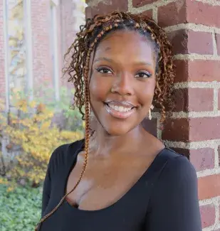 Headshot of Dejah Watts in a black shirt standing against a brick column