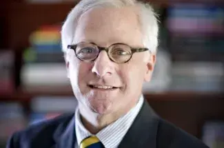 A close up bust of former Dean Michael Tick smiling at the viewer. A shelf with many colorful books is blurred in the background.