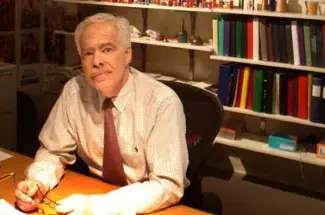 Color photo, Dean Robert Shay looks into the camera while he sits at a warm, dramatically lit desk with shelves of multi-colored binders and files lining the wall behind him.