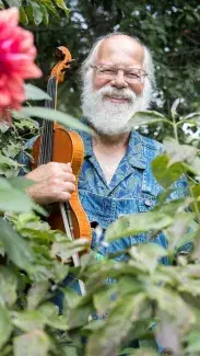 A happily grinning Ron Pen stands in a lush garden holding a fiddle close to his chest.
