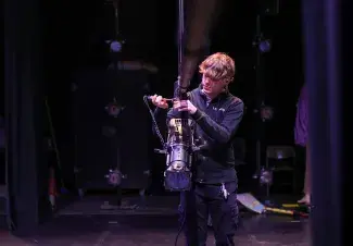 A student dressed in black connecting and securing a stage light on a lowered over-stage rig.