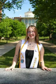 Jenna Perkovich stands center leaning against the polished granite sign for the W.T. Young Library which appears in the sunny distance behind her. She smiles happily, with honors cords and banner draped over her shoulders. Many verdant trees flank the paths that back to library.