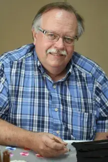 Bob Haven in a blue tartan button up shirt smiling at the camera while working on an embroidery project in a large frame.