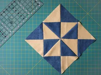 An assembled pinwheel block make of soft gold and denim blue fabric resting on a gridded cutting mat with a quilting ruler nearby.