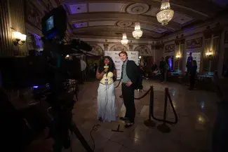 Two students stand in a historical theatre foyer with chandeliers and fancy architectural details. They are lit by several bright stage lights with a camer in the foreground recording them. One holds a mic while the other laughs. A film festival banner can be seen in the background.