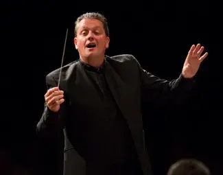 Bradley Genevro in a dark theatre and all black suit passionately conducting a band with a thin baton in one hand.