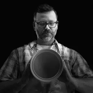 B/W headshot of Kyle Carpenter wearing a flannel shirt and glasses holding a piece of pottery up to the camera, Photo credit: Nancy Green