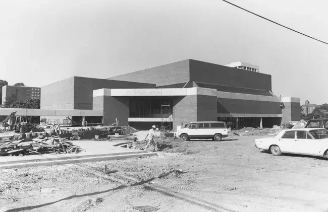 Black and white photo of the Center for the Arts under construction. The iconic silhouette of the building is complete but inside can be seen rebar and scaffolding. Several old cars, vans, and trucks are parked outside near piled construction materials. Most of the exterior is dirt with none of the broad walkways or grass lawn that exists today.