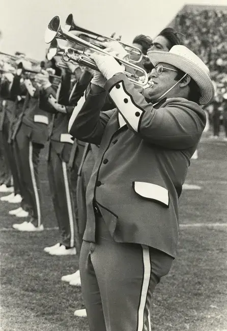 A black and white photo of the 1976 Marching Band plays in what is likely to be Commonwealth Stadium. A row of the brass section is lined up playing on the field. The band member in the foreground has dark slacks with white and black piping down the side of the leg, the jacket has white pocket flaps with lots of black piping along the hems and seams, white gloves, and a white fedora to keep the sun off his face.