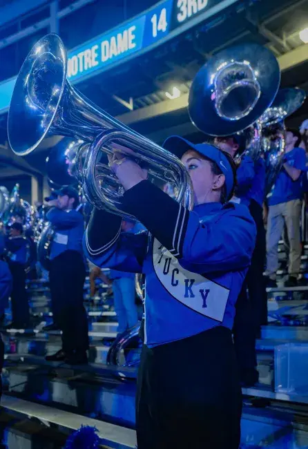 The Wildcat Marching Bands stands and plays in the bleachers of Kroger Field during a game with Notre Dame. Current uniforms have black pants, a UK blue and black jacket, with an embroidered KENTUCKY sash stitched across the front. Th band member in the foreground plays her silvery Euphonium, with other brass section members in the background playing a verity of instruments.