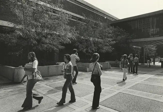 A black and white photo of the Margaret I King North Wing building in 1976. Several students walk in multiple directions on the broad sidewalk. The old pedway to the main King Library building can be seen in the far right background. Large planters along the face of the building contain trees that reach the second story.