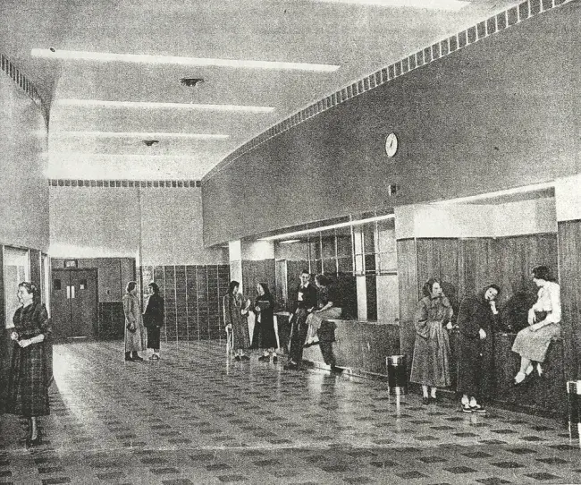 A scan of a black and white newspaper clipping showing the Guignol Lobby in 1952. The checkered tiling on the floor draws a straight line between front and inner doors. The lower walls around the whole lobby are a mixture of painted plaster, wood paneling, and ceramic tiles. A band of dark tiling circles the room where walls meet ceiling, and long strips of bright lights illuminate the space. Students gather and talk in various places around the lobby, possibly waiting for a performance.