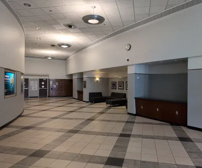 The current Guignol Lobby, a little empty as the school year ends. The tiling is now diagonal across the floor with three shades of gray akin to a muted simplified tartan. The lower half of the walls are now a uniform cool gray with a metallic strip highlighting the border with the lighter paint above. The star-studded box office desk remain, as well as some of the burgundy tiling next to the inner doors. The long and narrow lighting strips have been replaced with round pendant fixtures.