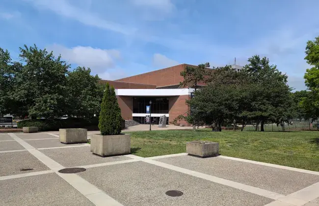 Singletary Center in May 2026. Many mature trees now obscure the building's silhouette, and the broad sidewalk is completed in the foreground. A blue sky with wispy white clouds frames the building from above.
