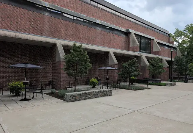 A color photo of the Fine Arts and Design Library building in 2026. Some tables with chairs as well as benches can be seen along the face of the building. A woman is reading some documents at one of the tables. Much newer trees are planted in smaller landscaping sections. A large floor to ceiling window can be seen on the second floor over the main entrance where the pedway used to be.