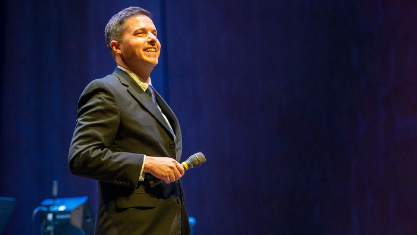 Image of Matt Gibson onstage at the Singletary Center wearing a suit holding a microphone